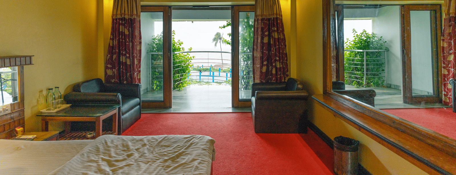 A bedroom with large windows leading to a balcony, 2 armchairs, a table, a mirror, wooden ceiling, and a king size bed at Hotel Seagull Digha.